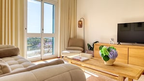 A modern living room with a sofa, a coffee table with a book and a vase of flowers, a television, and a balcony with curtains. - Funchal Infinity, a Home in Madeira (Funchal)