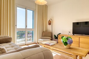 A modern living room with a sofa, a coffee table with a book and a vase of flowers, a television, and a balcony with curtains. - Funchal Infinity, a Home in Madeira (Funchal)