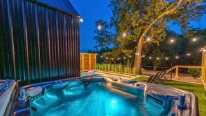 A hot tub with a wooden deck, surrounded by a fence and trees, under string lights. - A-Frame Retreat with Pool, Hot Tub, Lake Access (Garland County)