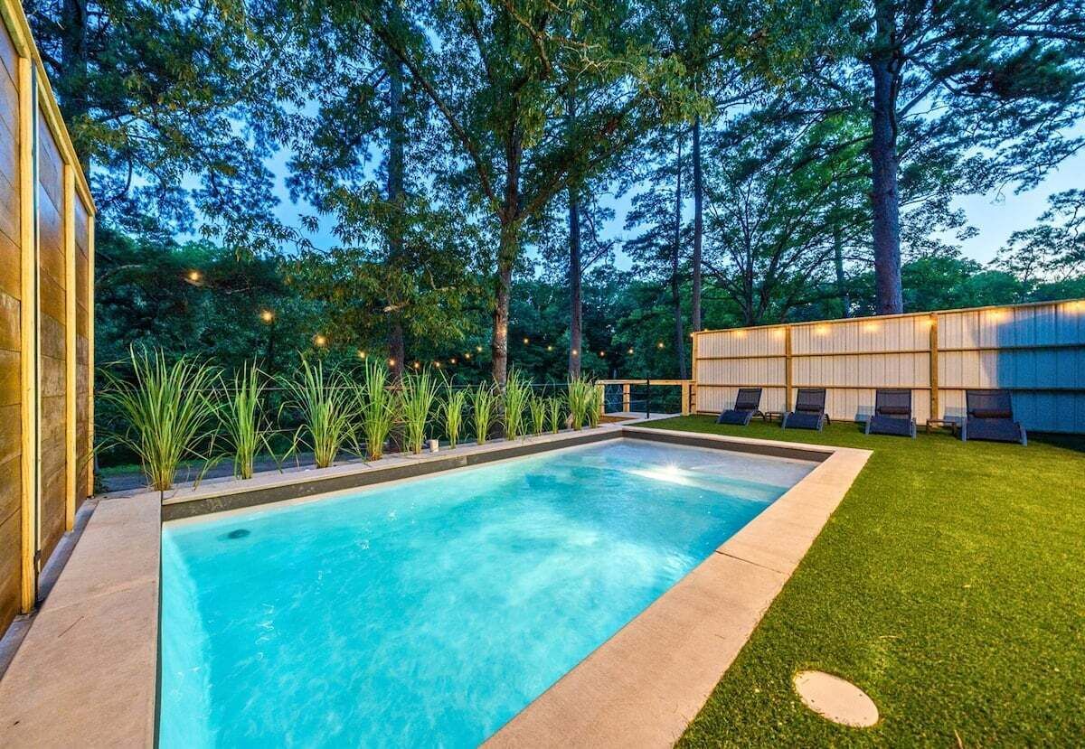 A rectangular swimming pool with a wooden deck, surrounded by a well-manicured lawn and tall trees.