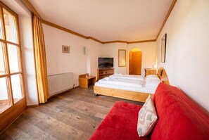 Habitación de hotel con cama, televisor, sofá rojo, ventana con cortinas y suelo de madera.