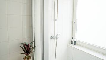 Un baño con azulejos blancos, ducha con cabezal de ducha manual, una planta en maceta y dos botellas en una estantería.