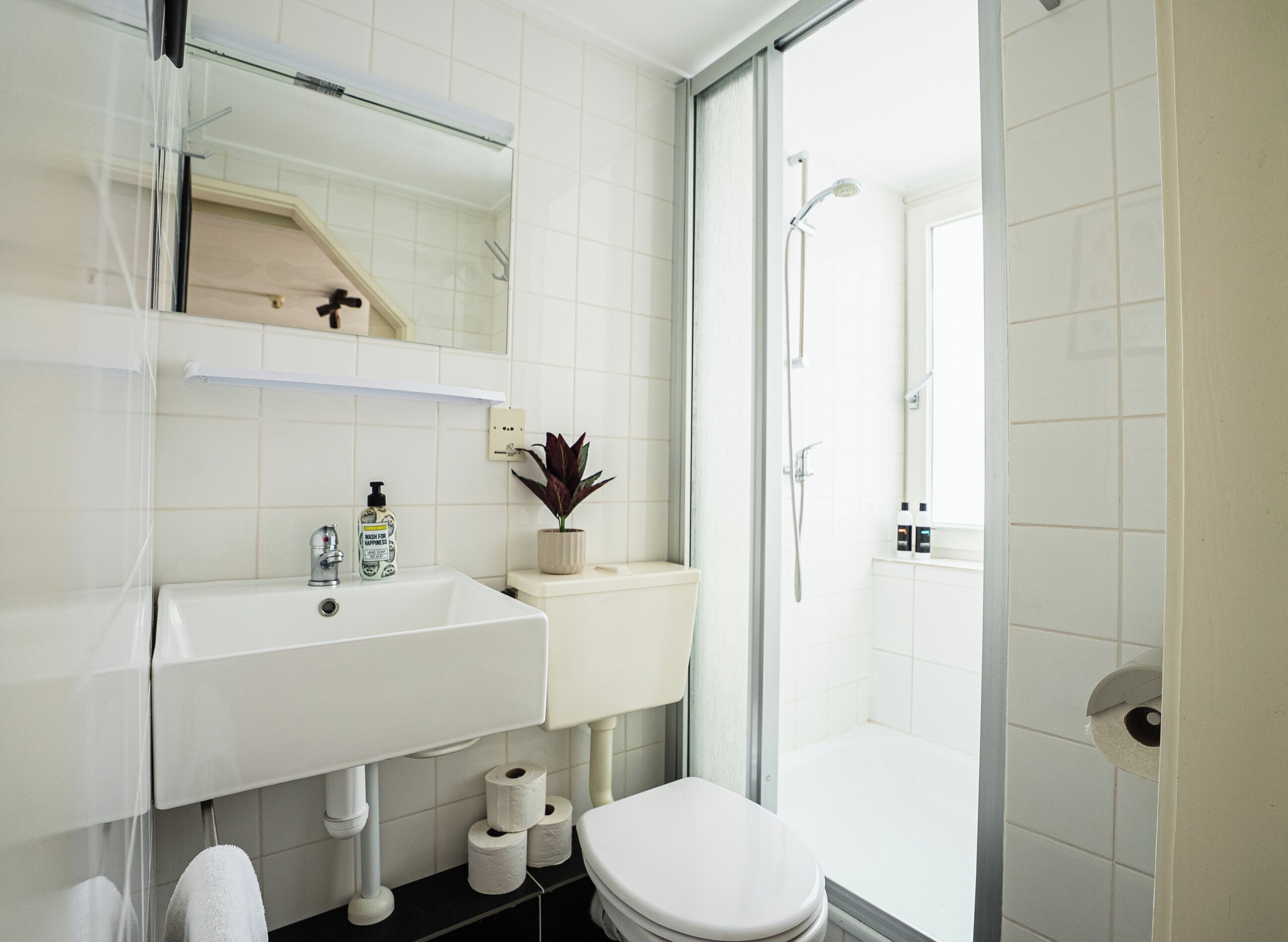 Un baño con lavamanos, inodoro y ducha blancos. Hay un espejo sobre el lavamanos y una planta en la pared.