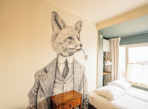 Ein Zimmer mit Wandgemälde eines Fuchses im Anzug, einem Holznachttisch, einem Bett mit weißer Bettwäsche und einem Fenster mit Vorhängen.