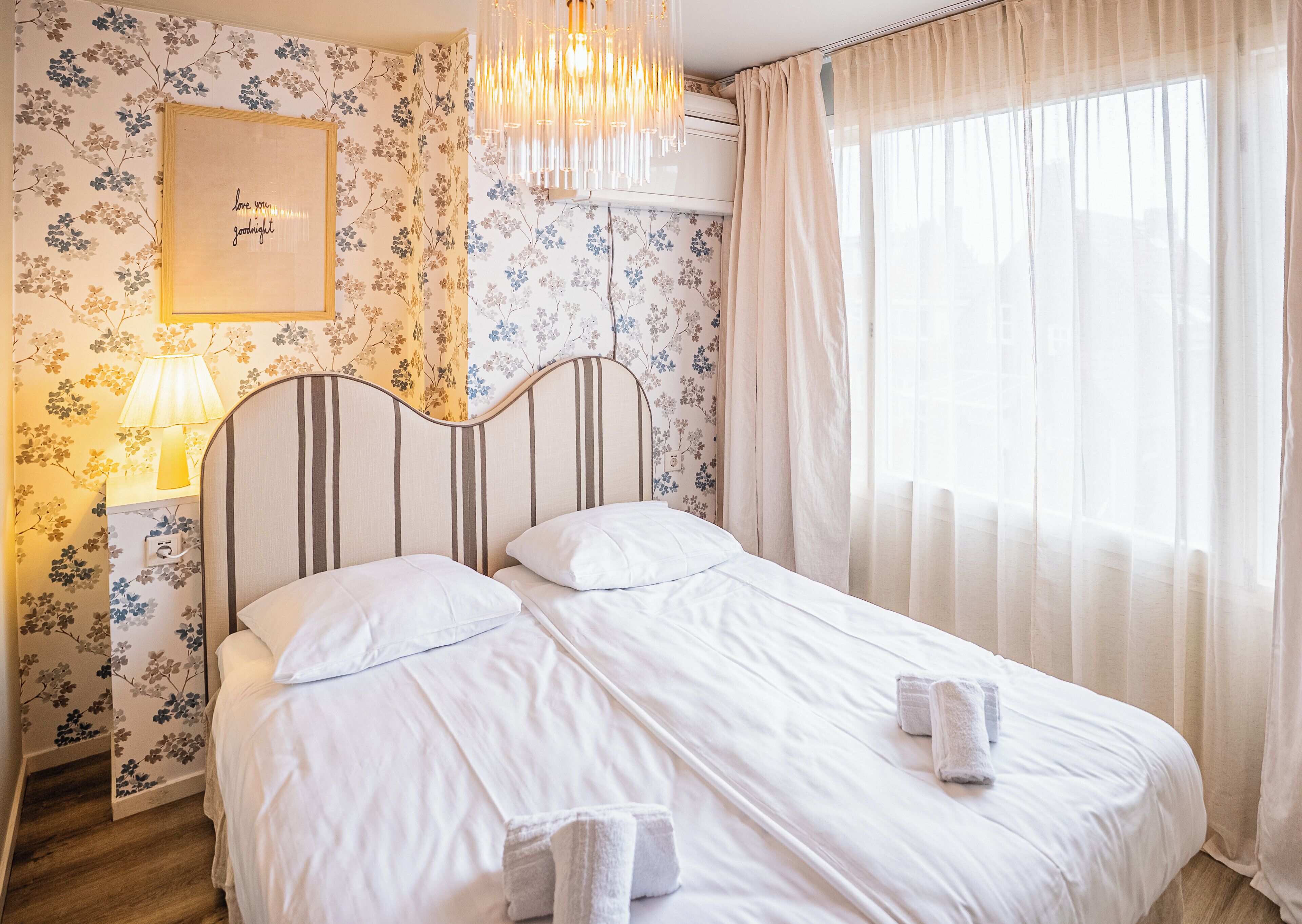 Una cama bien hecha con sábanas blancas, una lámpara de noche, un cartel en la pared y una ventana con cortinas traslúcidas.