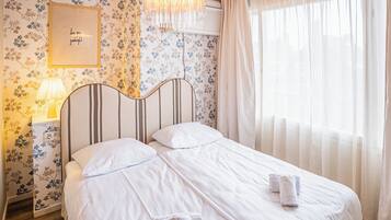 Una cama bien hecha con sábanas blancas, una lámpara de noche, un cartel en la pared y una ventana con cortinas traslúcidas.