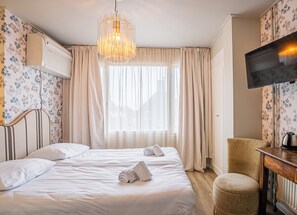 Ein Hotelzimmer mit einem Bett, zwei Kissen, einem Nachttisch mit Wasserkocher, einem Sessel, einem Fernseher und einem Fenster mit Vorhängen.