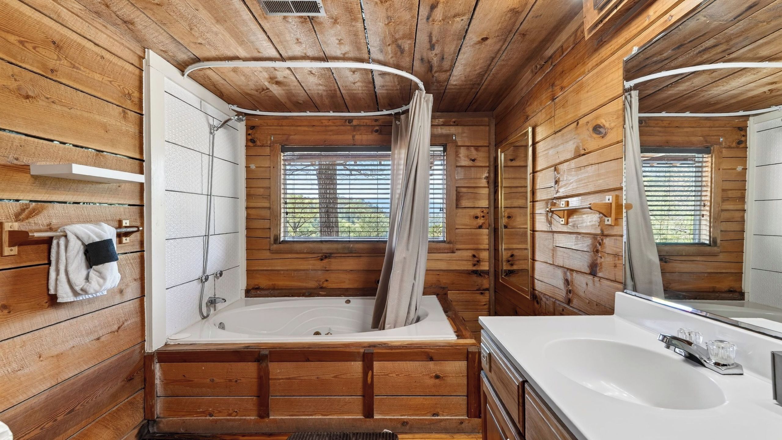 Baño de cabaña de madera con bañera, lavamanos, espejo y ventana con persianas.