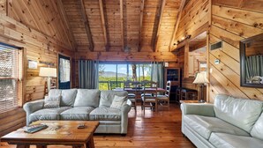 A cabin interior with wooden walls, a sofa, a dining table, and a view of the outdoors. - Elijah's, Isaiah's, Jack's & Sónyah's Cabins by Avantstay 4 Hot Tubs + Firepit Mountain Views + Decks 9min to Great Smoky Mtns National Park (Townsend)