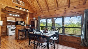 A wooden cabin interior with a dining area, kitchen, and large windows offering a view of the outdoors. - Elijah's, Isaiah's, Jack's & Sónyah's Cabins by Avantstay 4 Hot Tubs + Firepit Mountain Views + Decks 9min to Great Smoky Mtns National Park (Townsend)