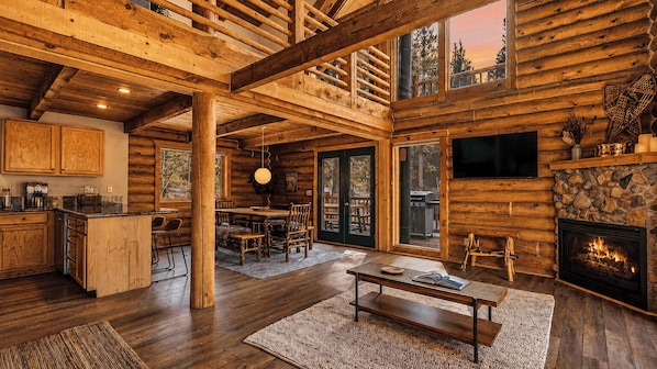 A log cabin interior with a dining area, kitchen, fireplace, and large windows.