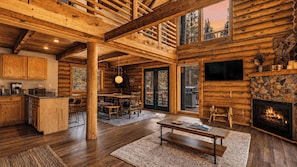 A log cabin interior with a dining area, kitchen, fireplace, and large windows.
