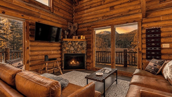 A log cabin interior with a fireplace, leather furniture, a television, and a view of a mountainous landscape.