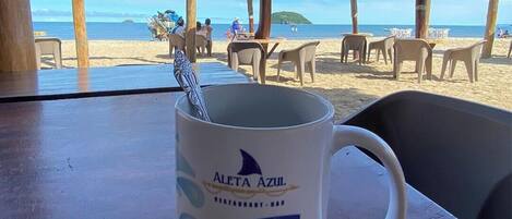 A beachside restaurant with a view of the ocean, tables with chairs, and thatched umbrellas.