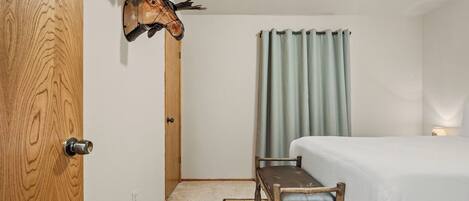 A room with a bed, a wooden chair, a wall-mounted animal head, and a curtain.