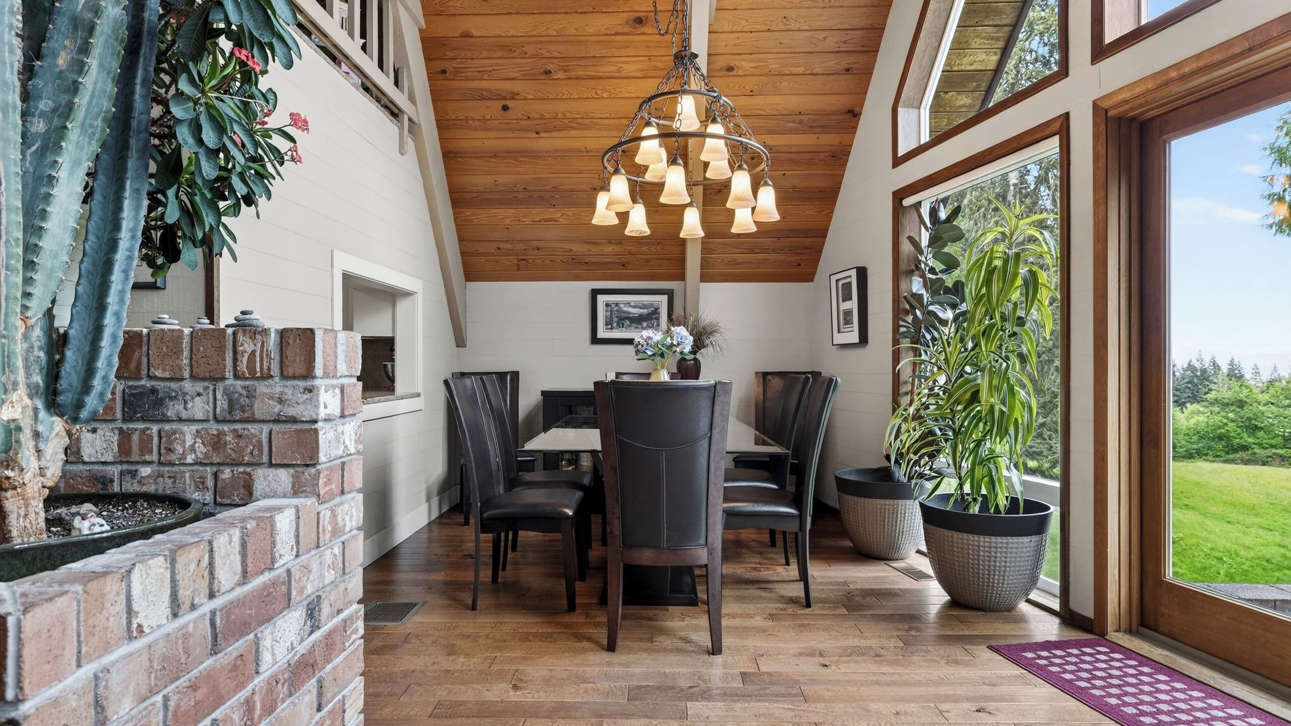 A dining area with a brick fireplace, wooden ceiling, and large windows overlooking a green landscape.