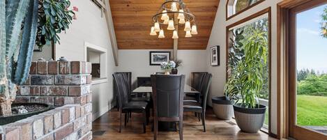 A dining area with a brick fireplace, wooden ceiling, and large windows overlooking a green landscape.