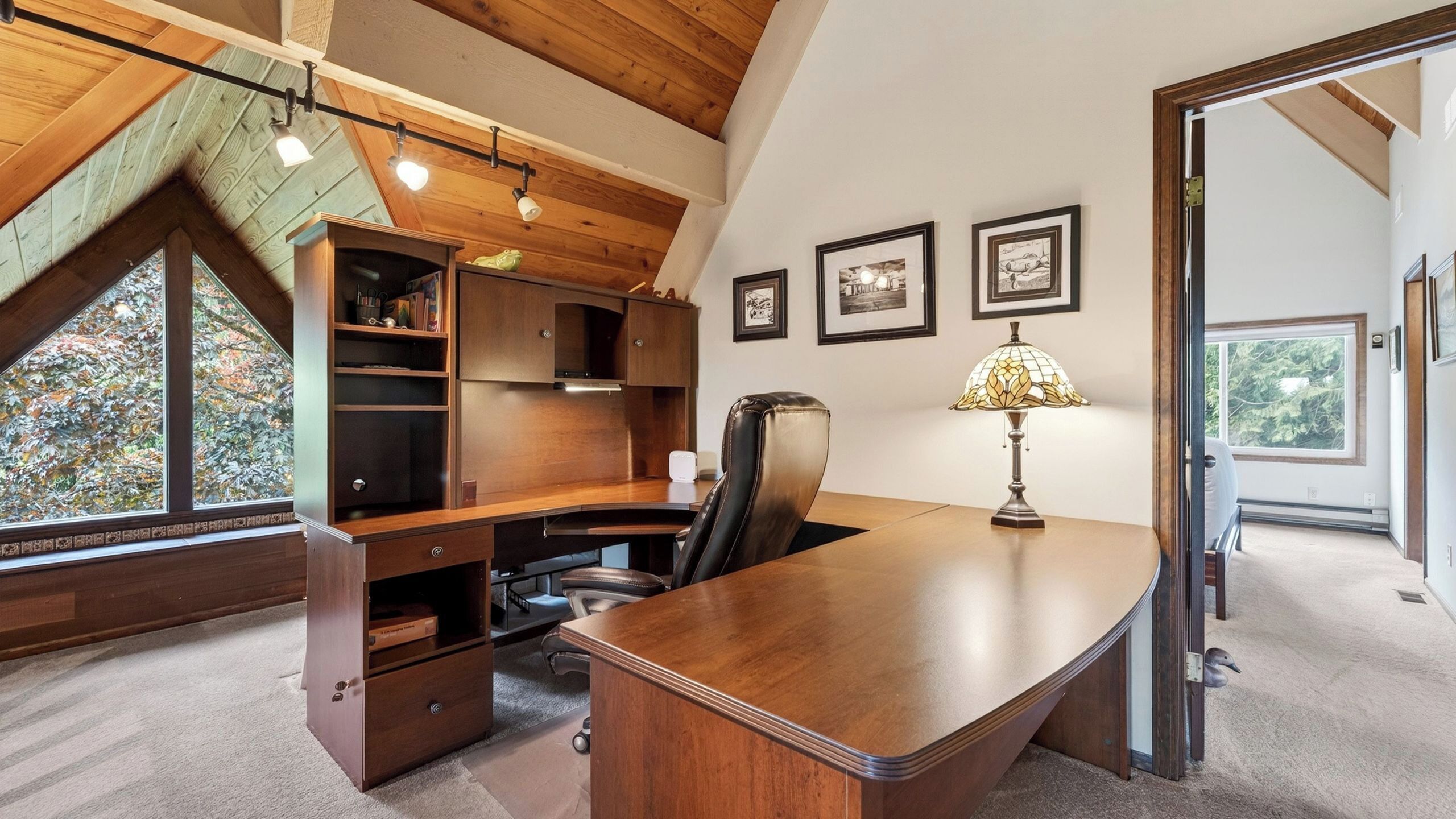 Een modern kantoor met een houten bureau, een lederen stoel en een lamp. Er hangen ingelijste foto's aan de muur en een raam met uitzicht op bomen.