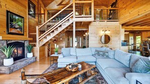 A log cabin interior with a stone fireplace, a large L-shaped sofa, a wooden coffee table, and a staircase leading to an upper level. - Serenity Ridge by Avantstay Spa + Game Room + Firepit Deck + Mountain Views Short Drive to Pigeon Forge (Sevierville)
