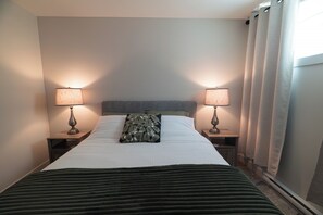 Una cama bien hecha con un cojín estampado, dos lámparas de noche y una ventana con cortinas.
