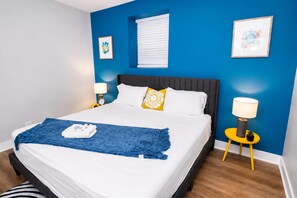 Quarto de hotel com cama, mesa de cabeceira, luminária e janela com persianas.