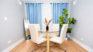 A modern dining area with a glass table, white chairs, blue curtains, and a potted plant. - Chicago Edgewater 3BR - 9 Min Walk to Loyola (Chicago)