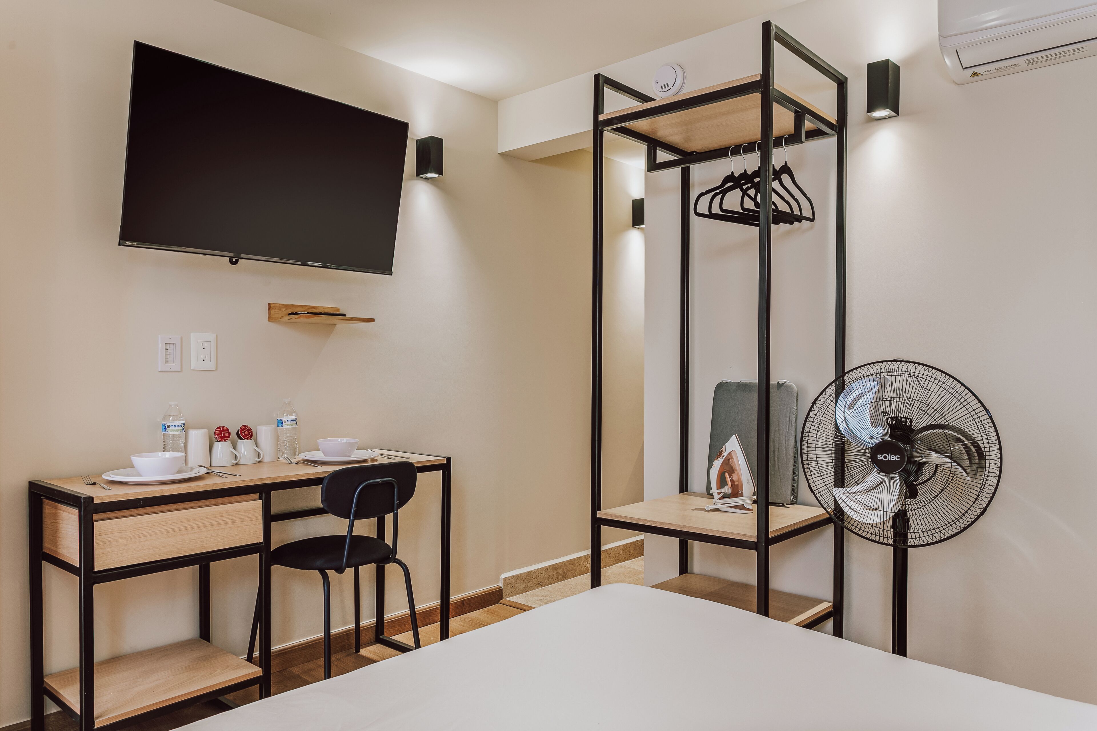 A hotel room with a bed, a desk with a chair, a TV mounted on the wall, a standing fan, and a wardrobe with a mirror.