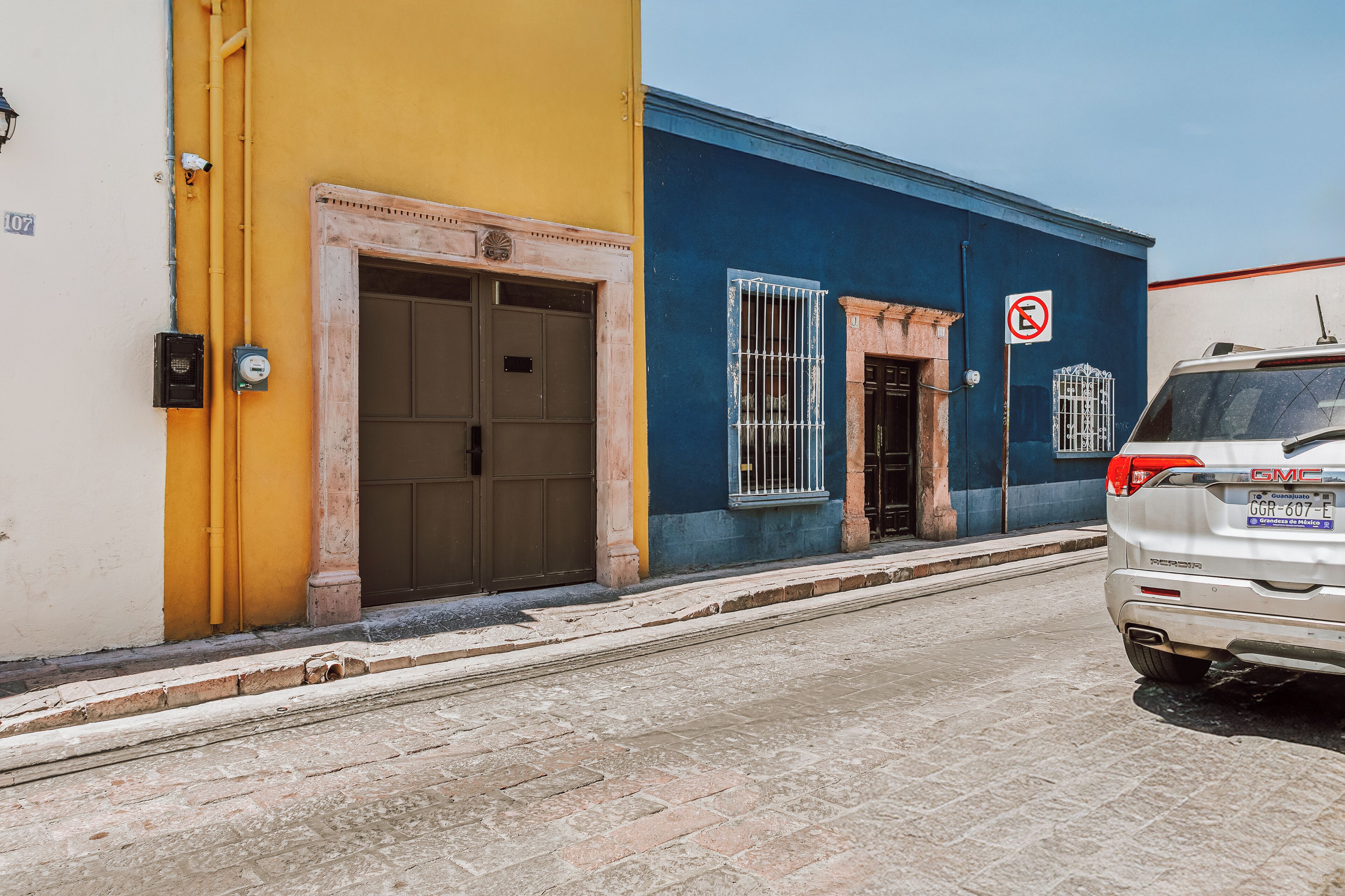 A street with a white GMC vehicle parked on the right, a building with a blue and yellow facade, and a no parking sign.
