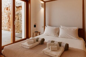 Ein Hotelzimmer mit einem großen Bett, einem Nachttisch, einer Lampe und Blick auf eine Steinmauer durch eine Glastür.