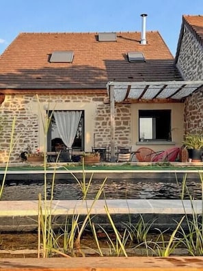 Ein Steinhaus mit einem Ziegeldach, einer kleinen Veranda mit Sitzgelegenheiten im Freien und einer Wasserspielszene im Vordergrund.