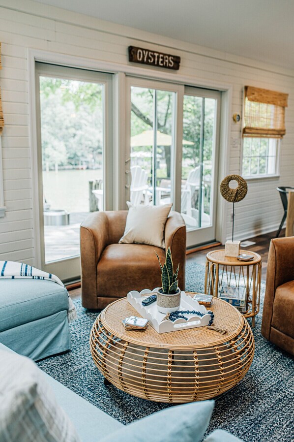 A cozy living room with a brown leather sofa, a round wicker coffee table with a plant and remote controls, and a sign above the door reading 'OYSTERS.'