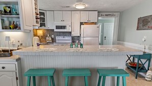 Una cucina con mobili bianchi, un frigorifero bianco, una cucina a gas bianca e uno sgabello da bar color petrolio.