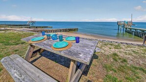 Un tavolo di legno apparecchiato con piatti e bicchieri su una spiaggia, con un molo sullo sfondo.