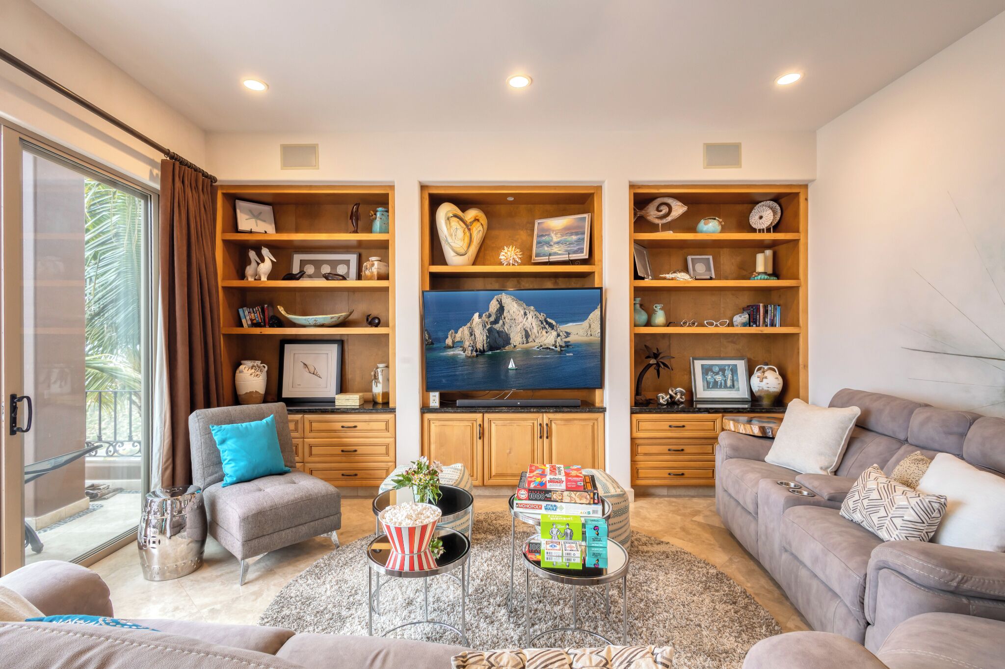 A living room with a sofa, armchairs, a coffee table, and a TV. There are shelves with decorative items and a painting on the wall.