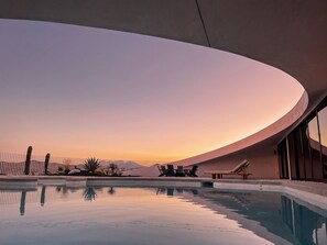 Ein moderner Poolbereich mit gewölbter Dachkonstruktion, Liegestühlen und freiem Blick auf den Sonnenuntergang.