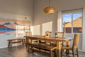 A dining area with a wooden table, benches, and chairs, a hanging light fixture, and a large wall mural. - Relaxing Desert Oasis · Hot Tub · Private Pool · 10 mins from National Park (Joshua Tree)