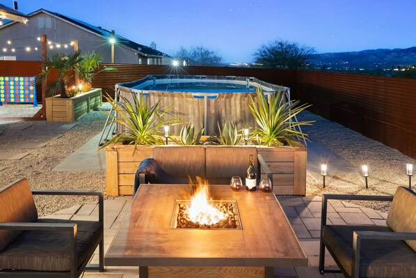 A backyard with a hot tub, a fire pit, outdoor seating, and a view of the city at night. - Relaxing Desert Oasis · Hot Tub · Private Pool · 10 mins from National Park (Joshua Tree)