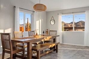 Un comedor con una mesa de madera, sillas, una lámpara colgante de fibra natural y vistas a montañas a través de amplios ventanales.