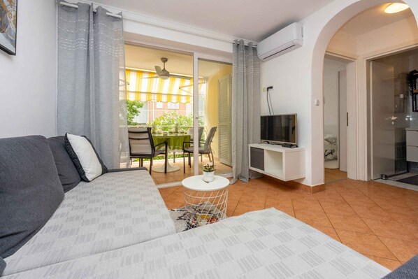 Ein Wohnzimmer mit einer Couch, einem kleinen Tisch, einem Fernseher auf einem Schrank und einer Glastür, die auf einen Balkon führt.