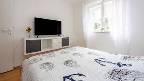 Una camera da letto con un letto, una TV su un mobile bianco, una finestra con tende trasparenti e un piccolo vaso con fiori.