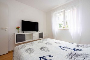Ein Schlafzimmer mit einem Bett, einem Fernseher auf einem weißen Schrank, einem Fenster mit durchsichtigen Vorhängen und einer kleinen Vase mit Blumen.