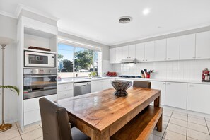 A modern kitchen with white cabinetry, a wooden dining table, and built-in appliances. - Sea View Steps to Central Esplanade Pet Friendly (Torquay)