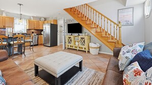 Una cocina moderna con armarios de madera, un comedor con mesa y sillas, una sala de estar con sofá y reposapiés, y una escalera de madera.