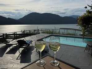 Zona de piscina con sillones de descanso y dos copas de vino blanco, con vistas a un lago y montañas al fondo.