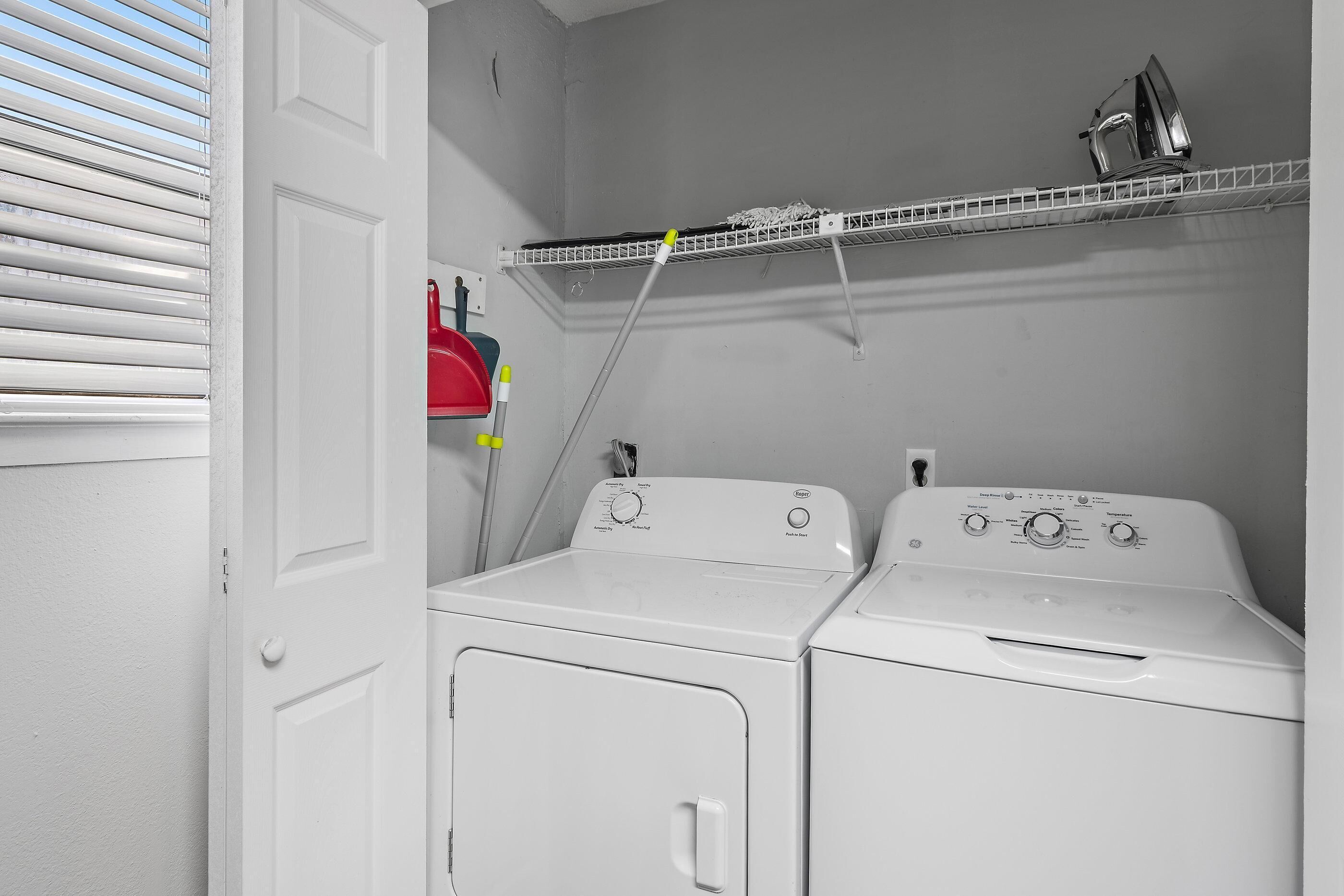 A laundry room with two white washers and dryers, a metal shelf above, and a red cleaning tool on the wall.