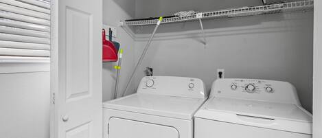 A laundry room with two white washers and dryers, a metal shelf above, and a red cleaning tool on the wall.
