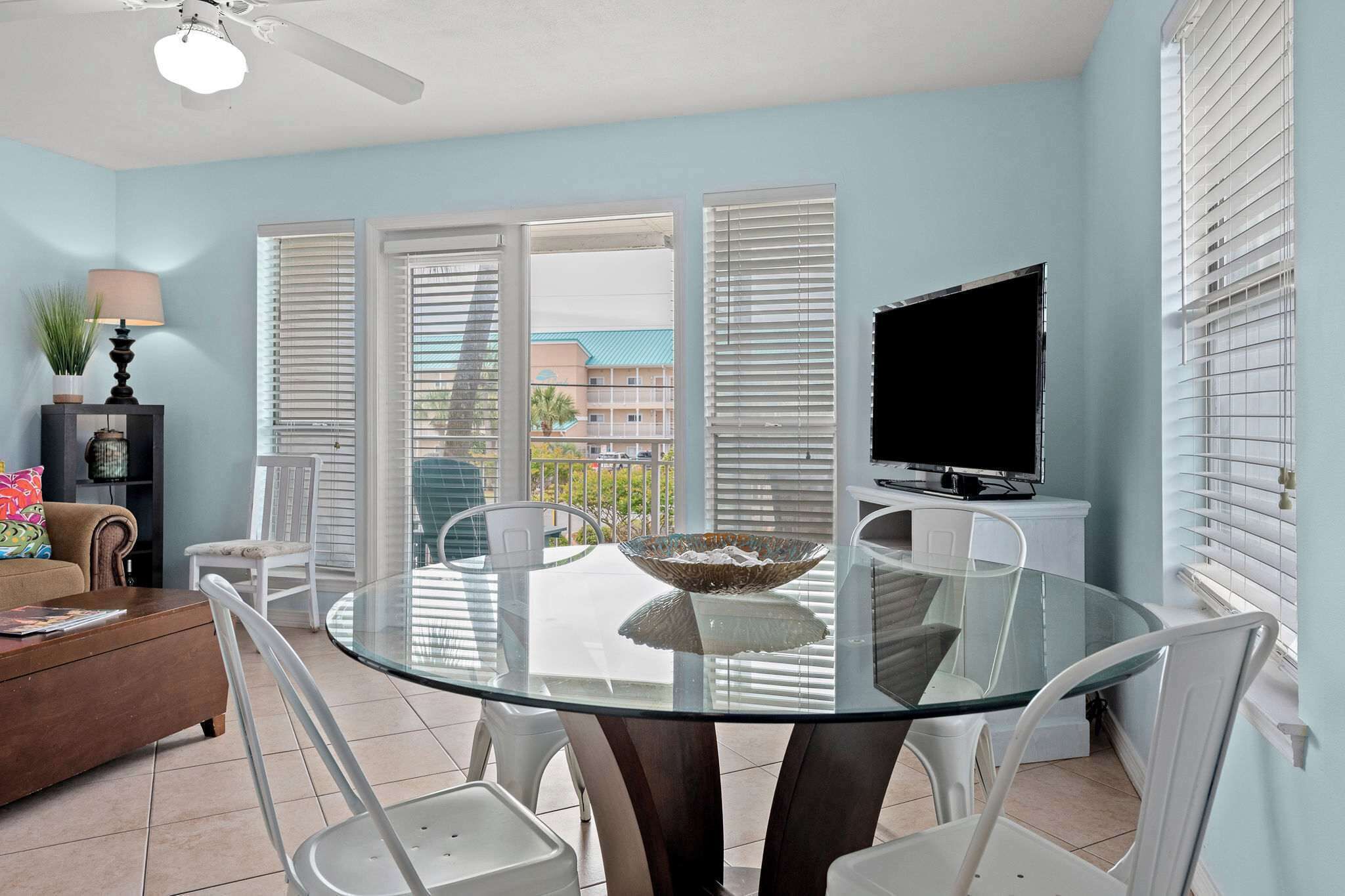 A dining area with a glass table, white chairs, a TV, and a view of buildings through open windows.