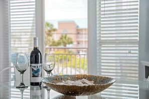 A bottle of wine, two wine glasses, and a decorative bowl on a table with a view of a balcony and buildings through the window blinds. - My SonShine-Hot Tub & Heated Pool-BeachView (Destin)