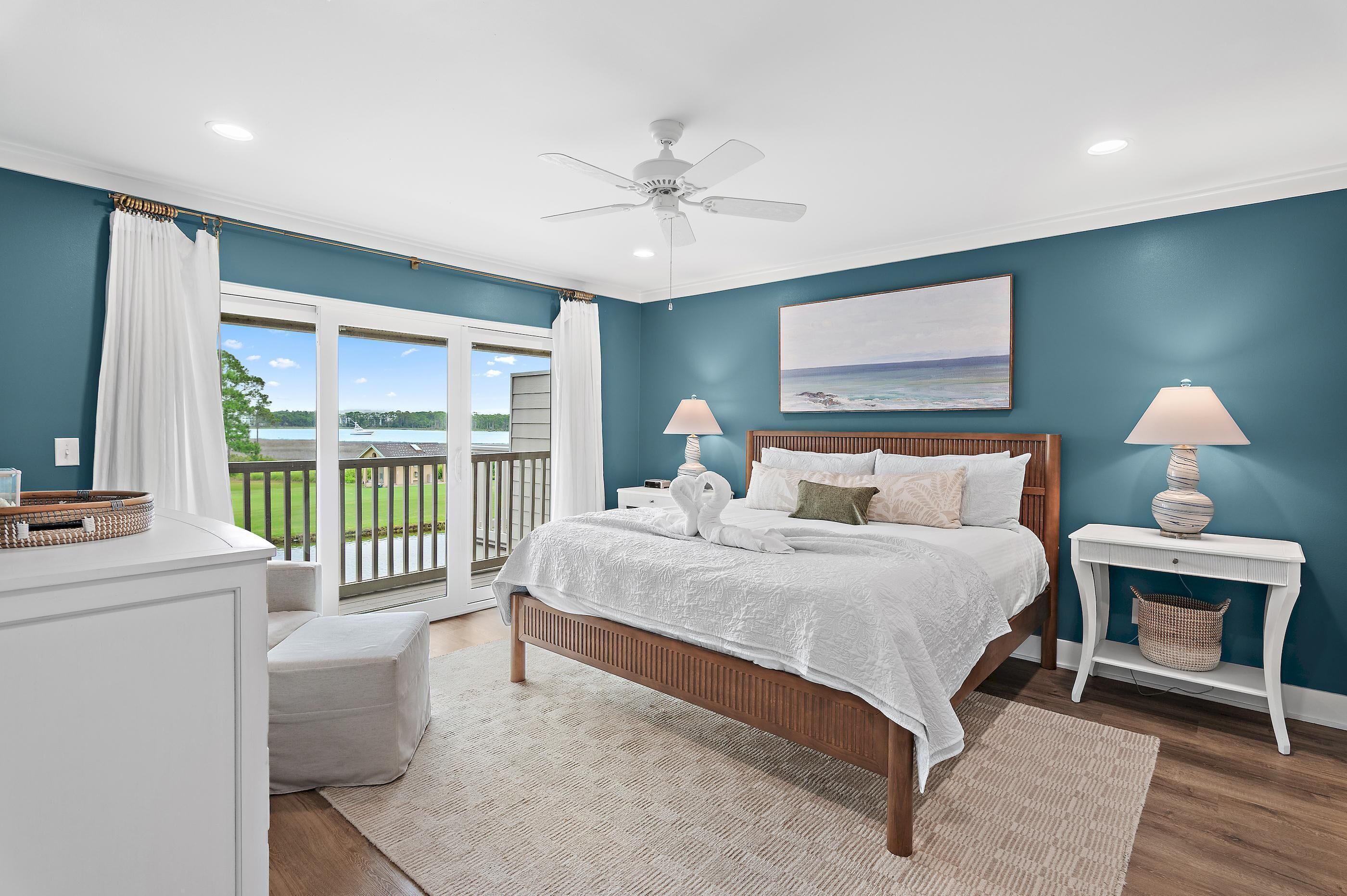 A bedroom with a bed, two bedside tables, a ceiling fan, a window with curtains, and a view of the outdoors.