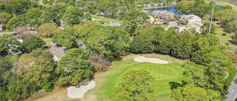 An aerial view of a golf course with sand bunkers, trees, and residential buildings in the background.